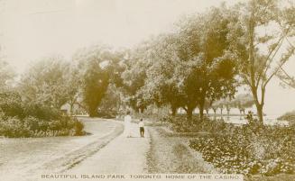Picture of two people walking on a path lined by large trees and gardens. 