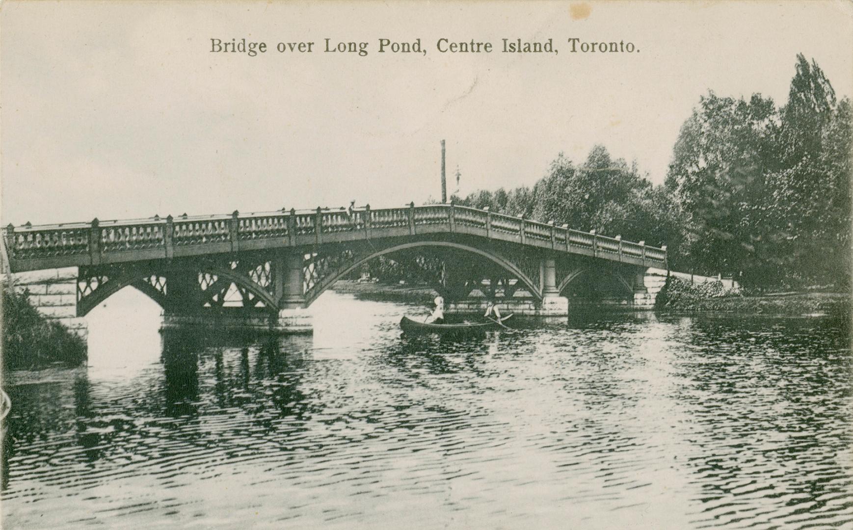 Black and white picture of two people in a canoe paddling under a bridge.