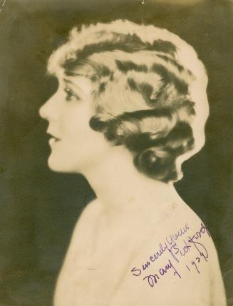 Profile portrait of Mary Pickford wearing short hair and a paste colored dress.