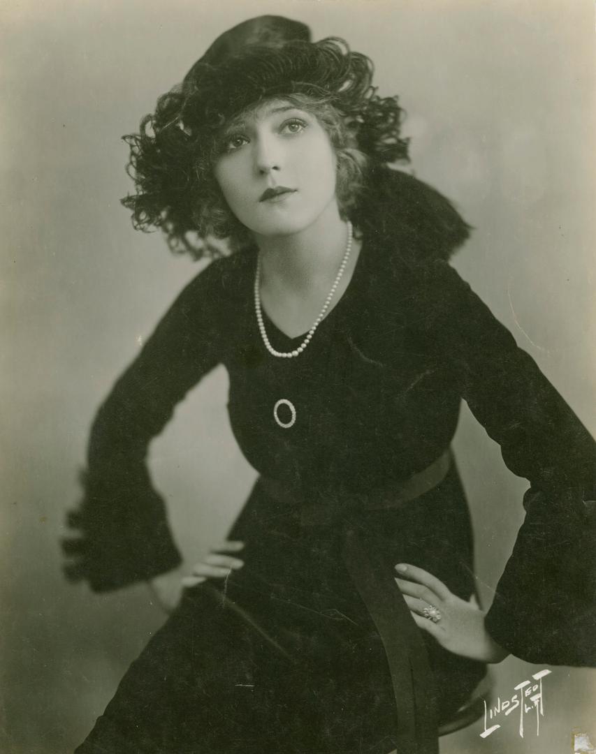 Portrait of Mary Pickford in a black coat and hat with her hands on her hips.