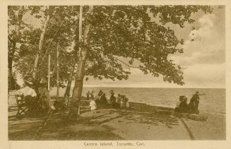 People in old fashioned clothing sitting and playing on a beach.