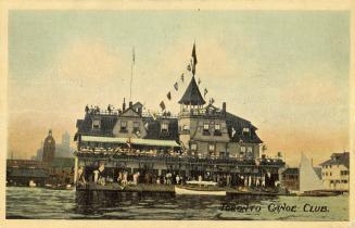 Colorized photograph of a large building beside a lake with many people standing on the dock in ...