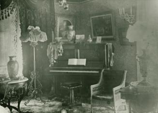 A photograph of an illustration of a room with a piano, chairs, lamp and other furniture.