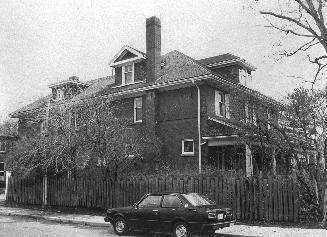 A photograph of a two-story house with a fence alongside it and a car parked in front of the fe ...