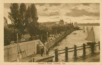 Black and white photo postcard of the boardwalk at Hanlan's Point, filled with people and showi ...