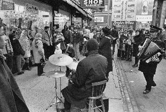 A group of musicians play for a crowd of people on the sidewalk outside of a store.