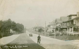 Black and white photograph of a police officer on a bicycle on a street with houses. Motor car …