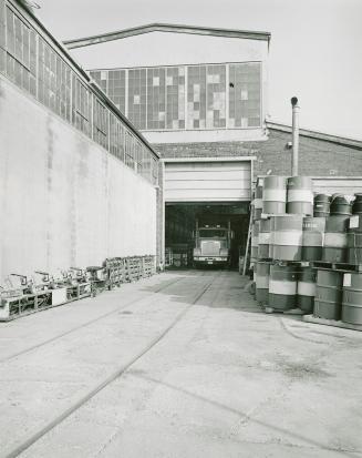 A photograph of a building with a transport truck parked inside an entranceway and barrels pile ...