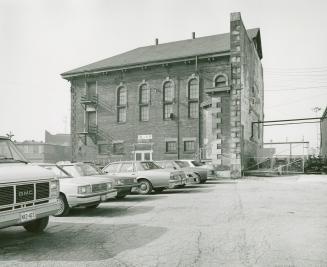 A photograph of a brick building with cars parked in a lot in front of it.