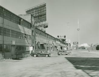 A photograph of a factory, with pickup trucks parked in a lot and the CN Tower visible in the b ...