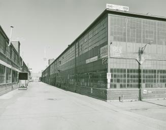 A photograph of two factory buildings, with an alleyway in between, and the CN Tower visible in…