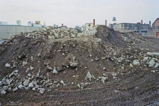A photograph of a pile of dirt and construction rubble in the middle of a muddy field, with hou ...