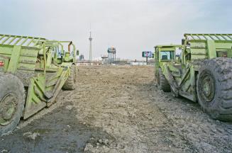 A photograph of two construction scrapers in a muddy field, with the CN Tower visible in the ba ...