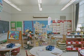 A photograph of a classroom, with desks, tables, shelves and easels, and assignments by student ...