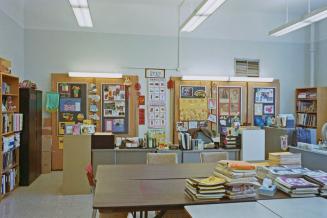 A photograph of a classroom, with desks, chairs, bookshelves and student assignments displayed  ...