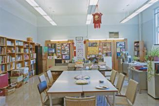 A photograph of a classroom, with tables, chairs, bookshelves and student assignments displayed ...
