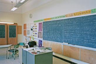 A photograph of a classroom, with a desk, chairs and a chalkboard.
