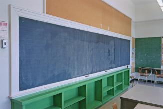 A photograph of a classroom, with tables, chairs, an empty shelving unit and a chalkboard.