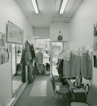 A photograph of a tailor's shop, with a chair, sewing station, racks of clothes and mirrors. 