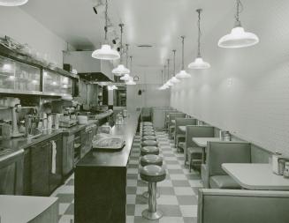 A photograph of a diner, with a kitchen area, counter, stools and tables visible. 
