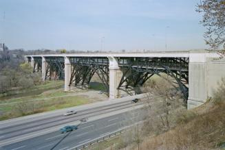 A photograph, facing west, of a bridge over the Don Valley in Toronto, with a highway visible u…