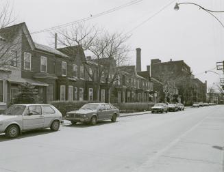 A photograph of a row of houses on the left side of a residential street, with cars parked on t ...