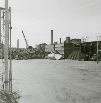 A photograph of an industrial plant area, with buildings, smokestacks and a crane.