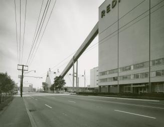 A photograph of a factory, with a chute leading from one building to another and a road in the  ...