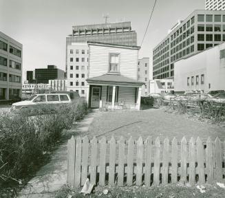 A photograph of a house, with a grass yard in front, surrounded on three sides by office buildi…