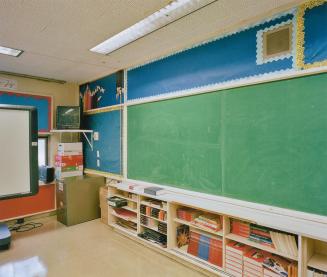 A photograph of a classroom, with a chalkboard, video screen and bookshelves. 