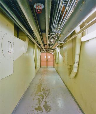 A photograph of a service corridor, with pipes overhead and a doorway at the end of the hall.