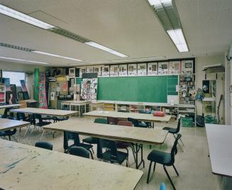 A photograph of a public school classroom, with chairs, tables, a bulletin board and artwork di ...