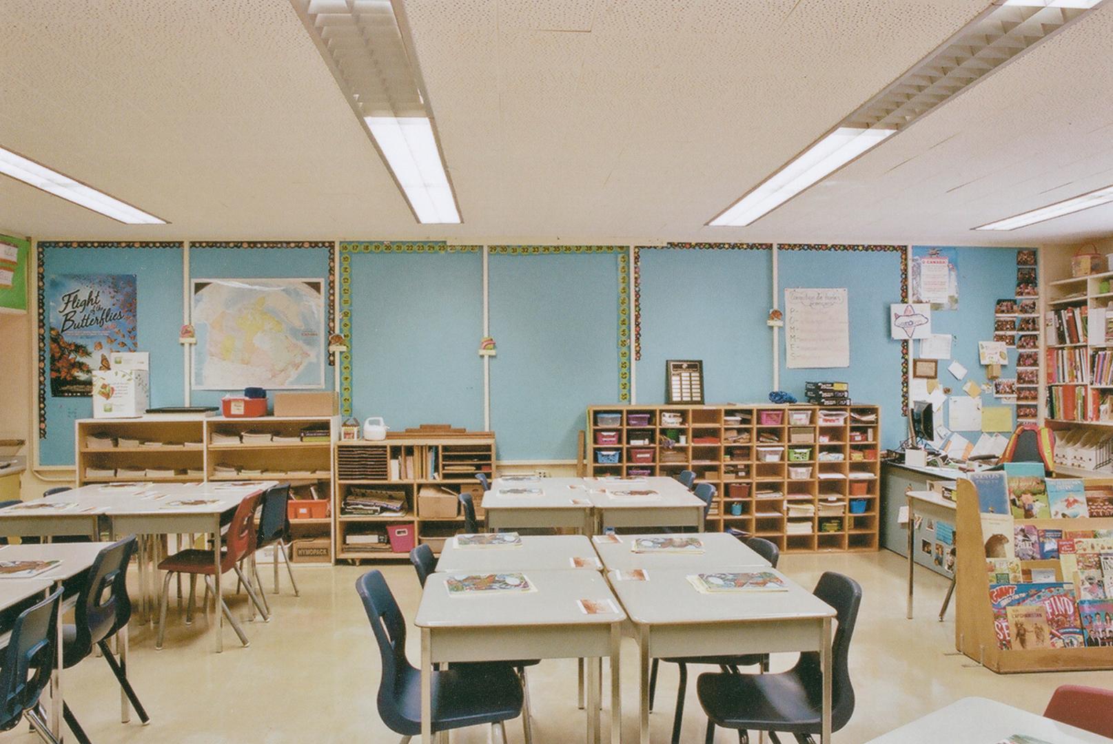 A photograph of a public school classroom, with desks, chairs, a chalkboard and shelving units.