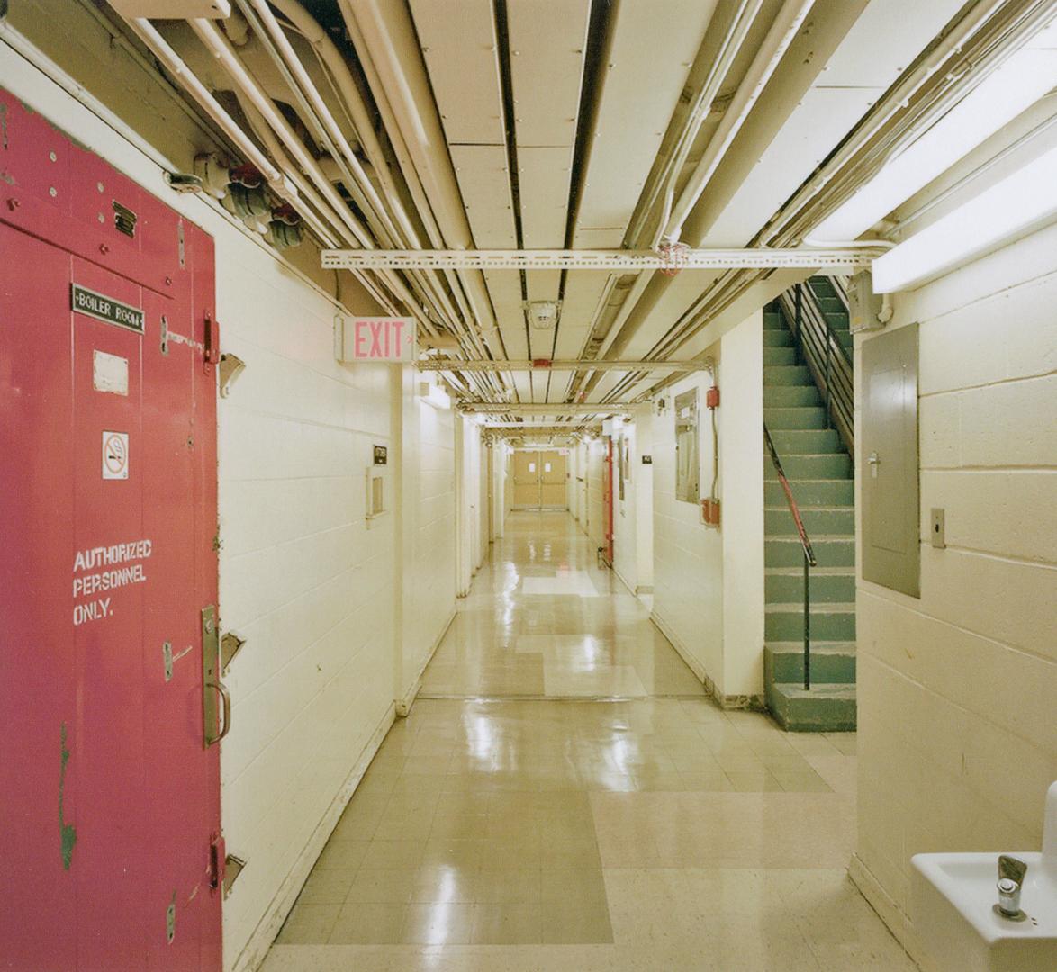 A photograph of a service hallway in the basement of a building with pipes overhead and a stair…