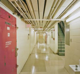 A photograph of a service hallway in the basement of a building with pipes overhead and a stair ...
