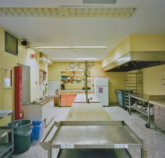A photograph of a cafeteria kitchen, with tables, a refrigerator and cupboards.