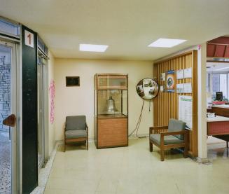 A photograph of an entrance foyer inside a public school, with a large bell inside a glass case ...
