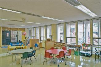A photograph of a kindergarten classroom, with round tables and chairs, and windows facing out  ...
