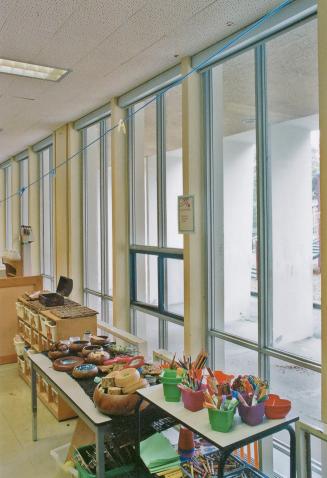 A photograph of a public school classroom, with tables and shelves in front of a wall of window ...