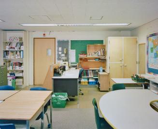 A photograph of a public school classroom, with chairs, tables, shelves and cabinets, a chalkbo ...