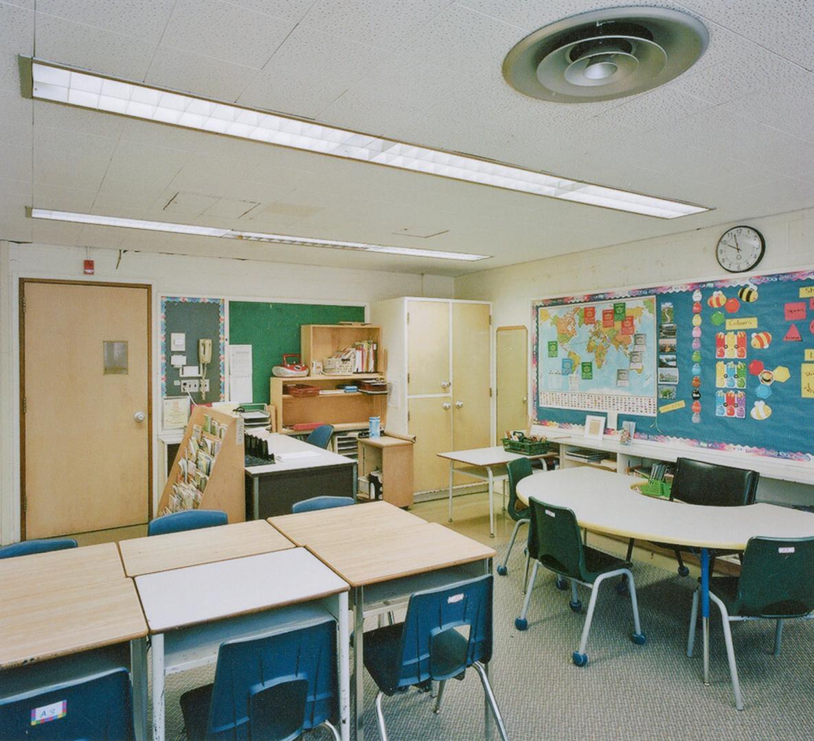 A photograph of a public school classroom, with chairs, tables, a chalkboard, shelves and cabin ...