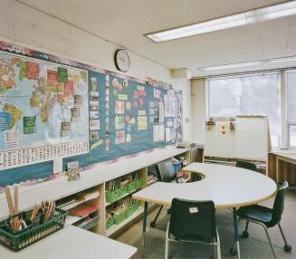 A photograph of a public school classroom, with chairs, desks, shelving units and a chalkboard.