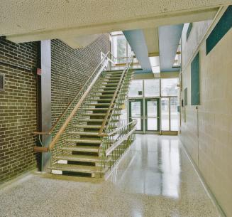 A photograph of a public school hallway, with a staircase rising to the second floor and window…