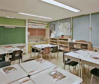 A photograph of a public school classroom, with chairs, desks, shelves and a chalkboard.