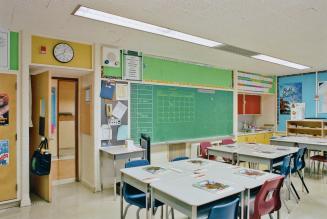 A photograph of a public school classroom, with chairs, tables, a chalkboard and a doorway open ...