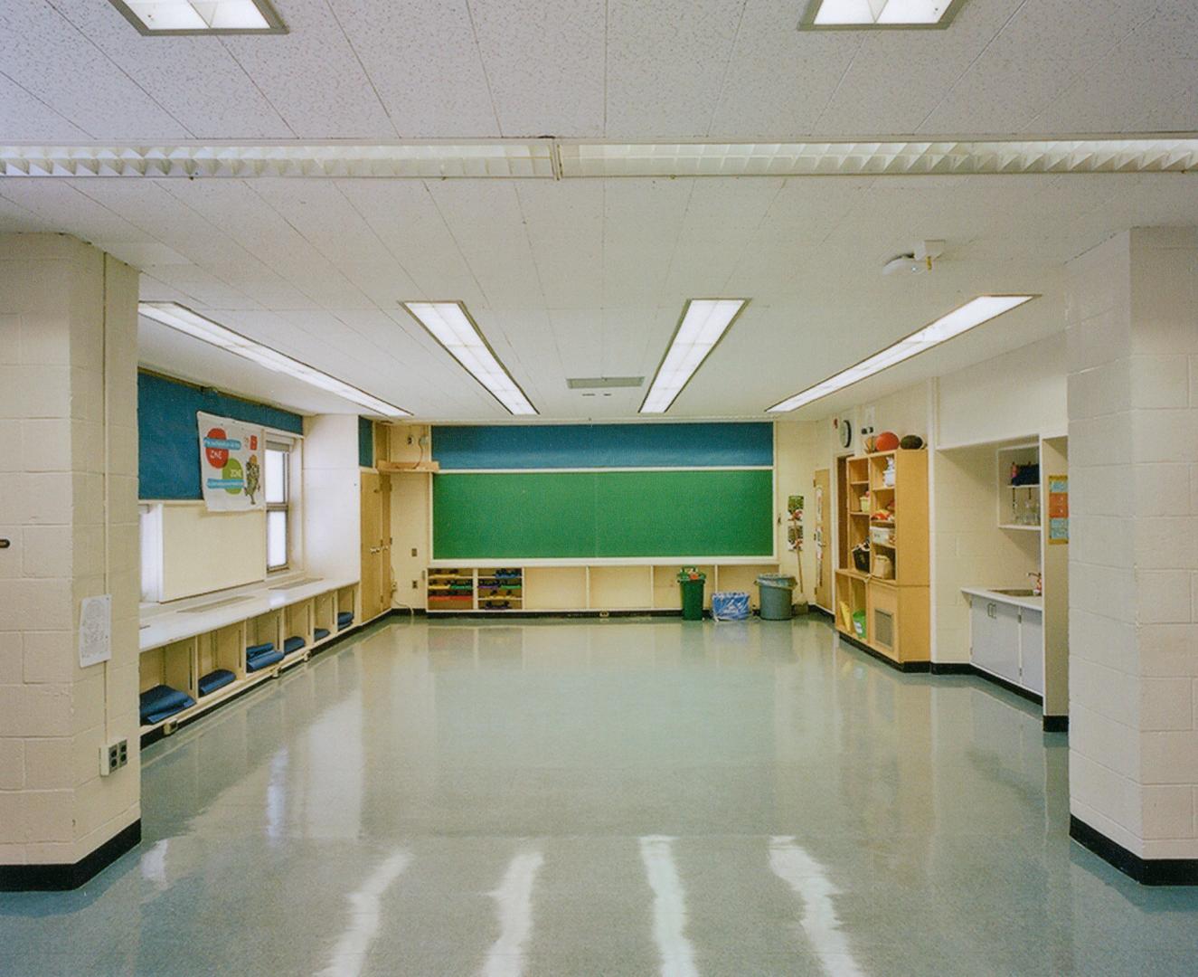 A photo of an open area in a public school, with a chalkboard, shelving units, a sink and windo ...