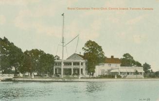 Colorized photograph of a large building with a veranda sitting at dockside with water in front…