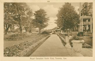 Sepia toned photograph of a walk way through gardens. Large building with veranda to the right.