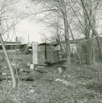 A photograph of a rudimentary wooden structure in a vacant lot, with buildings in the backgroun ...