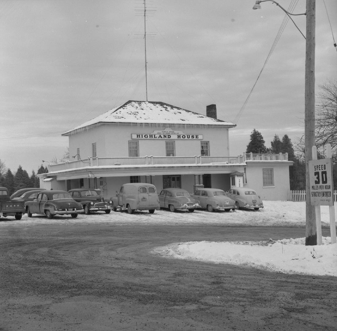 A photograph of a hotel with a sign reading &quot;HIGHLAND HOUSE,&quot; with cars parked in fro ...